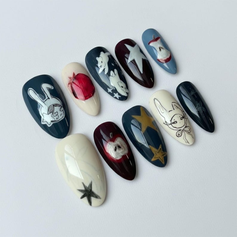 K&ouml;nnte beinhalten: Ein Set aus zehn k&uuml;nstlichen N&auml;geln mit verschiedenen Designs. Die N&auml;gel zeigen Illustrationen eines Kaninchens, Sterne und &Auml;pfel in Farben wie Marine, Wei&szlig;, Rot und Gold. Die N&auml;gel sind oval geformt und auf einer wei&szlig;en Oberfl&auml;che angeordnet.