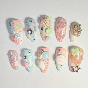 May include: A collection of ten almond-shaped, decorated artificial nails. Each nail features a unique design with pastel colors, including pink, blue, and yellow. Embellishments include buttons, stars, bows, and other decorative elements.
