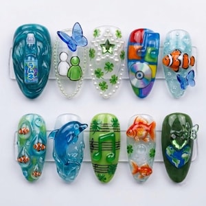 May include: A set of ten decorative press-on nails featuring 3D designs including a blue Ramune soda bottle, a blue butterfly, white and green figures, a star with pearls, clover patterns, a CD, clownfish, a blue dolphin, musical notes, and a globe.