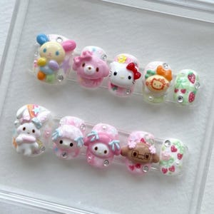 Peut inclure: Un ensemble de faux ongles avec des personnages de dessins animés et des motifs variés. Les ongles présentent une variété de couleurs, dont le rose, le blanc et des teintes pastel, avec des embellissements en 3D et des strass. Les motifs incluent un lapin, Hello Kitty et des fraises.