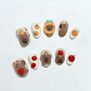 May include: A set of ten artificial nails with whimsical designs. The nails feature cartoon capybaras, oranges, and apples in various shades of brown, orange, and red, set against a white background. The nail art is detailed and playful.