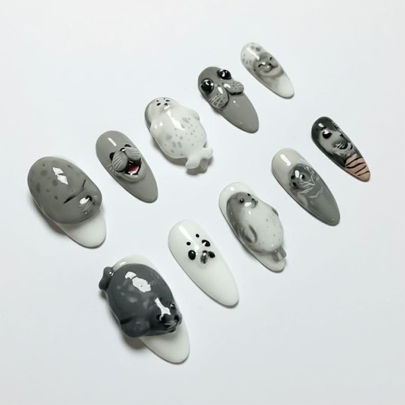 Puede incluir: Una colecci&oacute;n de diez u&ntilde;as artificiales, cada una con un dise&ntilde;o &uacute;nico de foca en 3D. Las u&ntilde;as presentan una variedad de colores, incluyendo blanco, gris y negro, con caras y cuerpos de foca detallados. El arte de u&ntilde;as muestra un tema l&uacute;dico y art&iacute;stico.