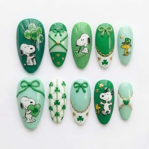 Könnte beinhalten: Ein Set aus zehn oval geformten Kunstnägeln in Grün- und Hellgrüntönen mit verschiedenen Designs. Die Designs zeigen Snoopy mit Ballons und einem Herz, Kleeblätter, Schleifen und Woodstock. Einige Nägel haben goldene Akzente.