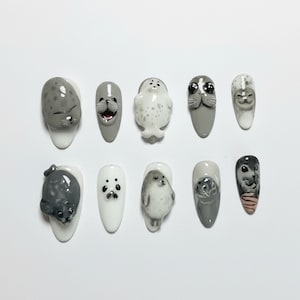 Puede incluir: Diez dise&ntilde;os de u&ntilde;as con focas. Las u&ntilde;as est&aacute;n pintadas en varios tonos de gris y blanco, con caras y cuerpos de foca detallados esculpidos en cada u&ntilde;a. Los dise&ntilde;os incluyen diferentes poses y expresiones, creando una est&eacute;tica l&uacute;dica.