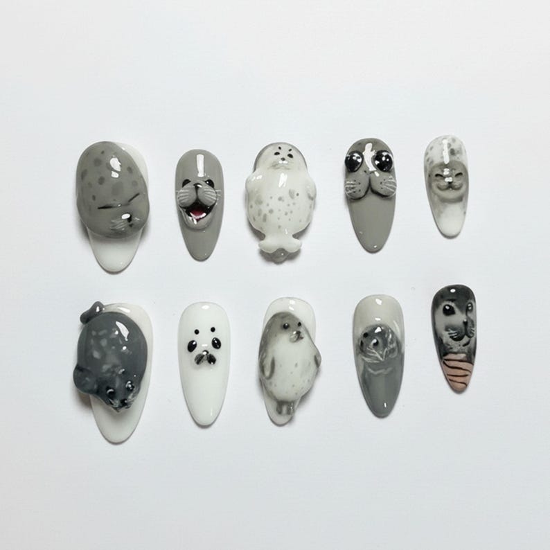 Puede incluir: Diez dise&ntilde;os de u&ntilde;as con focas. Las u&ntilde;as est&aacute;n pintadas en varios tonos de gris y blanco, con caras y cuerpos de foca detallados esculpidos en cada u&ntilde;a. Los dise&ntilde;os incluyen diferentes poses y expresiones, creando una est&eacute;tica l&uacute;dica.