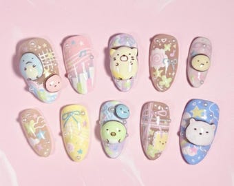 Può includere: Dieci unghie finte, ognuna con un design unico con colori pastello e personaggi dei cartoni animati. I disegni includono un orso, un gatto e altri simpatici personaggi, con elementi decorativi come fiocchi e stelle. Le unghie sono disposte su una superficie rosa.