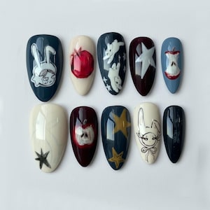 K&ouml;nnte beinhalten: Ein Set von zehn mandelf&ouml;rmigen Kunstn&auml;geln mit handgemalten Designs. Die Designs umfassen einen roten Apfel, einen wei&szlig;en Stern, ein Kaninchen und einen Stern. Die N&auml;gel sind in verschiedenen Farben wie Marineblau, Burgunderrot und Creme bemalt.