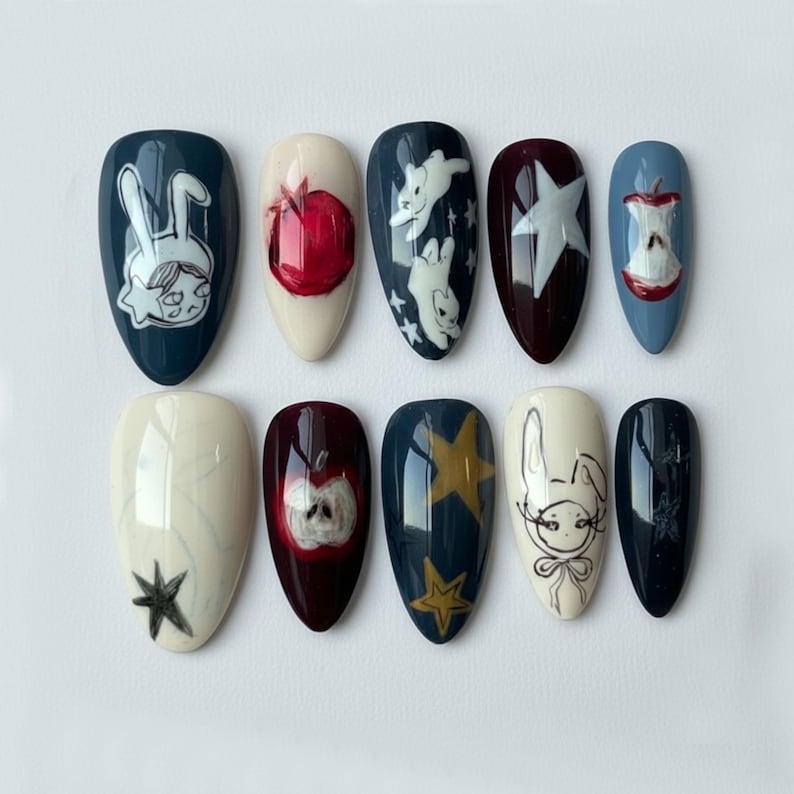 K&ouml;nnte beinhalten: Ein Set von zehn mandelf&ouml;rmigen Kunstn&auml;geln mit handgemalten Designs. Die Designs umfassen einen roten Apfel, einen wei&szlig;en Stern, ein Kaninchen und einen Stern. Die N&auml;gel sind in verschiedenen Farben wie Marineblau, Burgunderrot und Creme bemalt.