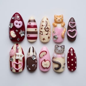 May include: A set of ten almond-shaped press-on nails featuring diverse designs including a burgundy base with heart buttons and stars, striped patterns, musical notes, 3D cat figures, apple slices, piano keys, and polka dots with rhinestones.