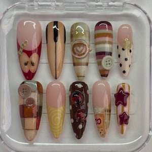 May include: A set of ten almond-shaped press-on nails in a clear plastic case. Designs include apple motifs, piano keys, a latte art charm, buttons, polka dots, swirls, ladybugs, and raised star accents in a palette of red, brown, tan, and cream.