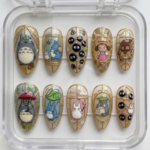 Könnte beinhalten: Ein Set aus zehn mandelförmigen Press-on-Nägeln mit handgemalten Illustrationen animierter Figuren wie Totoro, Rußmännchen und einem Katzenbus, akzentuiert durch goldene Linien, die an Buntglasrahmen erinnern, in einem durchsichtigen Etui.