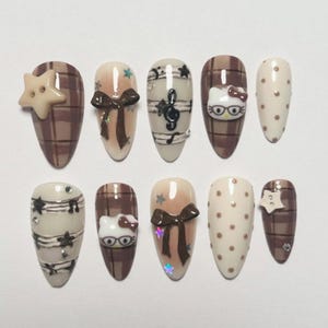 May include: A set of ten almond-shaped false nails with various designs. The nails feature brown plaid, musical notes, bows, polka dots, and a Hello Kitty design. Some nails have star and gem accents. The color palette includes brown, beige, and white.
