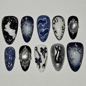 May include: A set of ten almond-shaped press-on nails featuring a dark color palette of black, navy, and white. Designs include white rabbit silhouettes, silver metallic charms, ornate frames, bows, cutlery, and celestial star patterns.