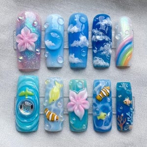 May include: A collection of ten artificial nails with various designs. The designs include a pink flower, clouds, a rainbow, and aquatic themes with fish and coral. The nails feature a glossy finish with glitter and 3D elements.