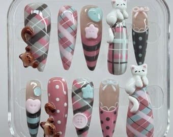 May include: A set of ten press-on nails in a clear plastic case, featuring various designs including pink and grey plaid patterns, polka dots, and solid colors. Several nails are adorned with 3D charms, including white cats, brown bears, and buttons.