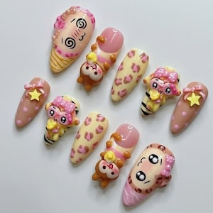 Op de afbeelding: Een set van tien kunstnagels met diverse ontwerpen, waaronder beige nagels met roze stippen en gele sterren, luipaardprints en 3D-karakterdecoraties met roze strikjes, gestreepte lichamen en expressieve cartoongezichten.
