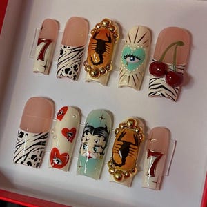 May include: A set of ten long, square-shaped artificial nails with various designs. The designs include a number 7, zebra print, a scorpion, a heart with an eye, cherries, and a Betty Boop illustration. The nails are in a white box.