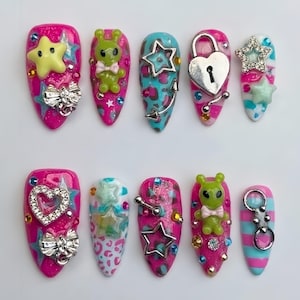 May include: A set of ten press-on nails featuring vibrant pink and blue designs with various 3D embellishments including green alien figures, silver heart-shaped padlocks, metallic star charms, small bows, rhinestones, and colorful leopard print patterns.