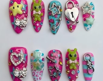 Puede incluir: Un juego de diez uñas postizas con diseños vibrantes en rosa y azul, decoradas con adornos en 3D que incluyen figuras de alienígenas verdes, candados plateados en forma de corazón, estrellas metálicas, lazos y brillantes de colores.