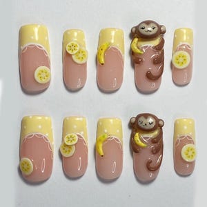 May include: A set of ten press-on nails with a fun design. Each nail has a light pink base, a yellow tip with white polka dots, and a white scalloped edge. Some nails feature banana or lemon slice embellishments, while others have a 3D monkey.