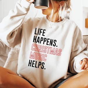 May include: A cream-colored sweatshirt with the text "LIFE HAPPENS. CHRISTMAS MUSIC HELPS." in black and red. The person is holding a black mug. The person is sitting on a gray couch.