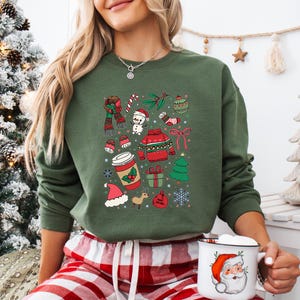 May include: An olive green crewneck sweatshirt featuring a colorful graphic print of various Christmas icons including a snowman, a red sweater, a coffee cup, a gift box, a Santa hat, and snowflakes. A white mug with a Santa face is also shown.