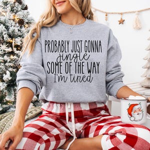 May include: A light gray sweatshirt with the text "Probably just gonna jingle some of the way I'm tired" in black. The person is wearing red and white plaid pajama pants and holding a Santa mug. A Christmas tree is in the background.