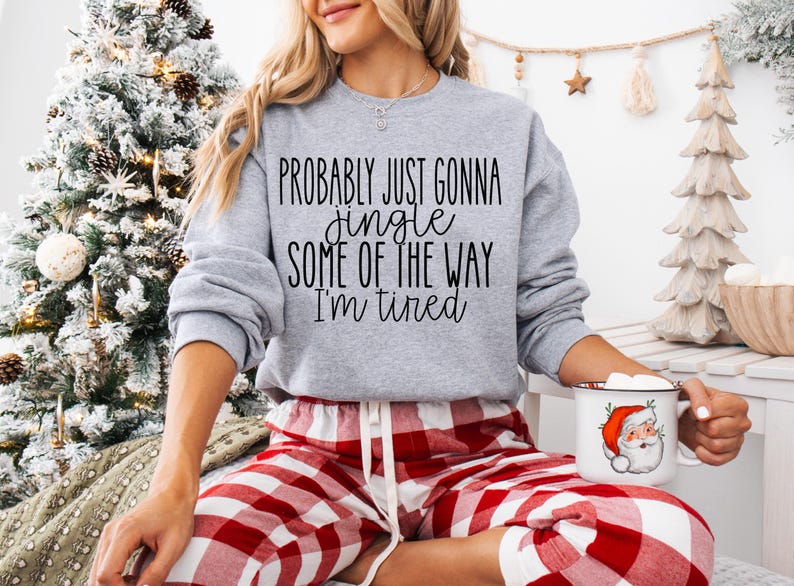 May include: A light gray sweatshirt with the text "Probably just gonna jingle some of the way I'm tired" in black. The person is wearing red and white plaid pajama pants and holding a Santa mug. A Christmas tree is in the background.