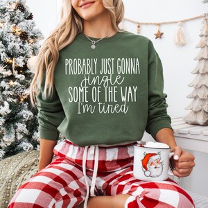 May include: A green sweatshirt with the text "Probably Just Gonna Jingle Some of the Way I'm Tired" in white. The person is wearing red and white plaid pajama pants and holding a mug with a Santa Claus graphic. A Christmas tree is in the background.