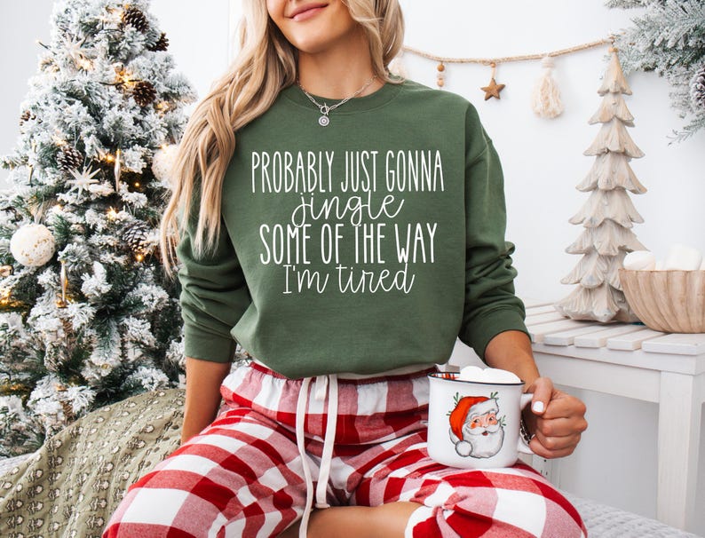 May include: A green sweatshirt with the text "Probably Just Gonna Jingle Some of the Way I'm Tired" in white. The person is wearing red and white plaid pajama pants and holding a mug with a Santa Claus graphic. A Christmas tree is in the background.