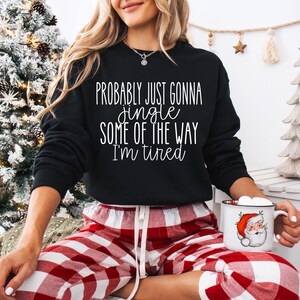May include: Black sweatshirt with the text "Probably just gonna jingle some of the way I'm tired." The person is wearing red and white plaid pajama pants and holding a mug with a Santa Claus graphic.