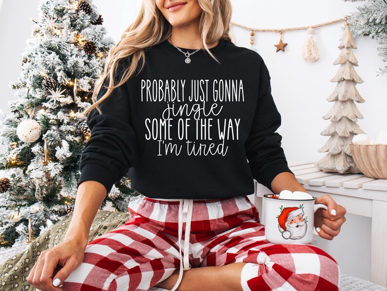 May include: Black sweatshirt with the text "Probably just gonna jingle some of the way I'm tired." The person is wearing red and white plaid pajama pants and holding a mug with a Santa Claus graphic.