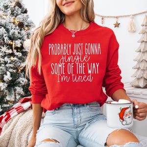 May include: A red sweatshirt with the text "Probably Just Gonna Jingle Some of the Way I'm Tired" in white script. The person is holding a white mug with a Santa Claus graphic. The sweatshirt is paired with light blue jeans.