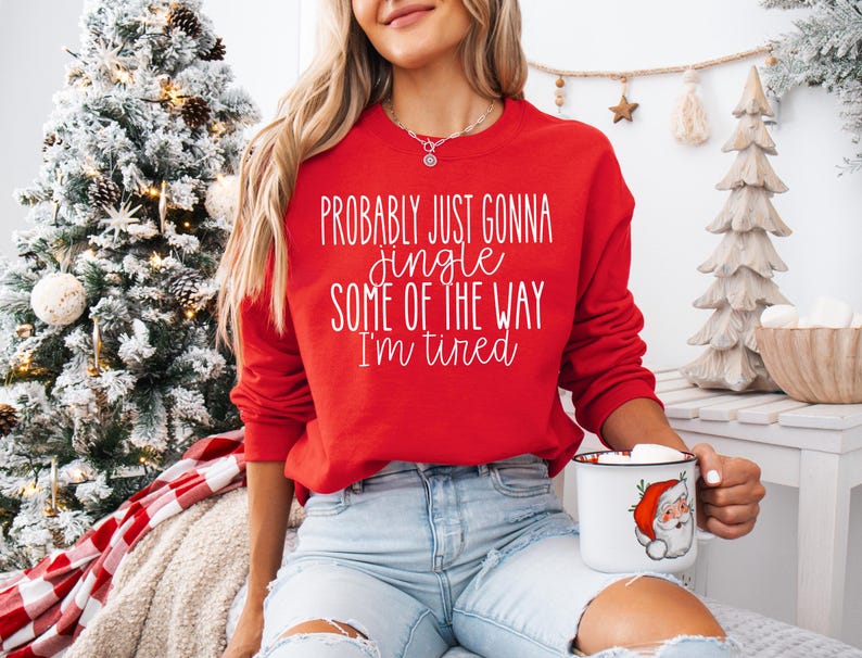 May include: A red sweatshirt with the text "Probably Just Gonna Jingle Some of the Way I'm Tired" in white script. The person is holding a white mug with a Santa Claus graphic. The sweatshirt is paired with light blue jeans.