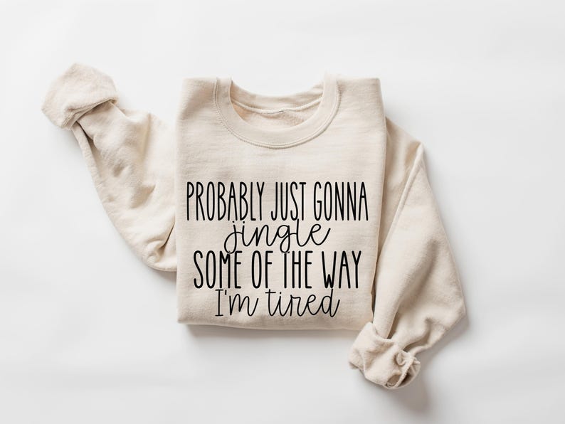 May include: A cream-colored sweatshirt with the text "Probably just gonna jingle some of the way I'm tired" in black script. The sleeves are folded, and the garment is laid flat on a white surface.