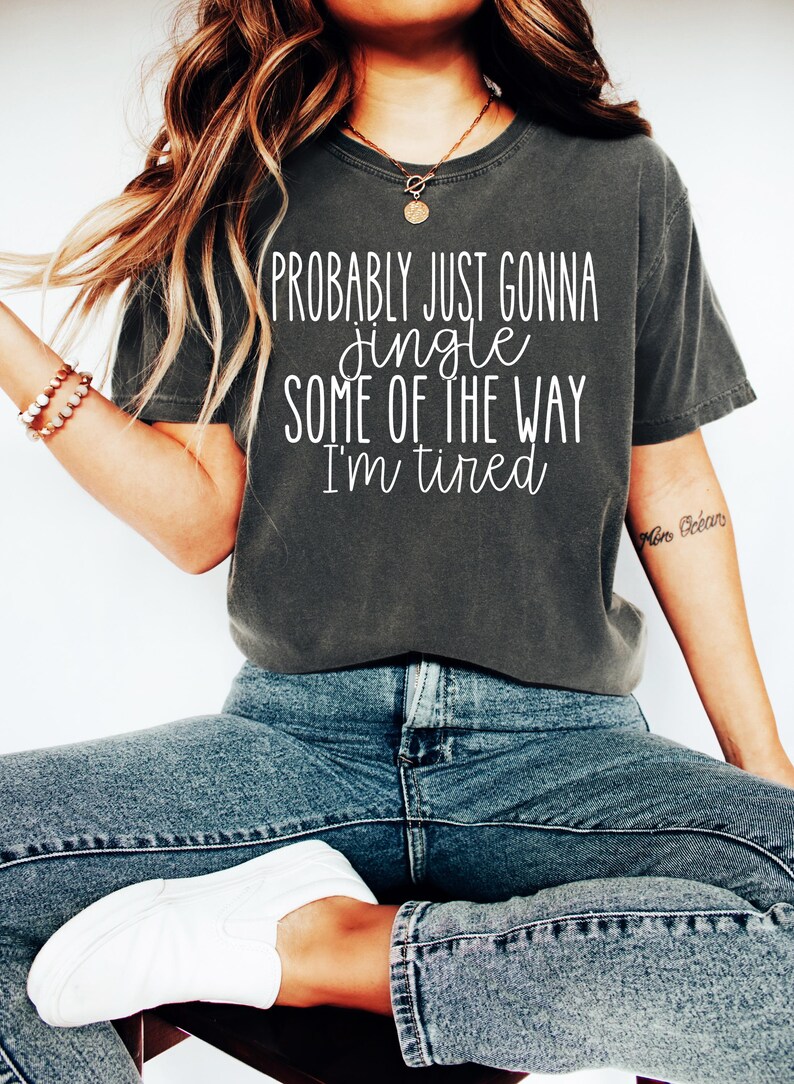 May include: Dark gray t-shirt with the text "PROBABLY JUST GONNA jingle SOME OF THE WAY I'M tired" in white. The shirt is paired with blue jeans and white sneakers. The overall style is casual and comfortable.