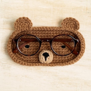 Puede incluir: Un soporte para gafas con forma de oso tejido a ganchillo de color marrón con dos orejas pequeñas y una nariz beige. El soporte contiene un par de gafas redondas con montura de carey. El fondo es una superficie de madera clara.