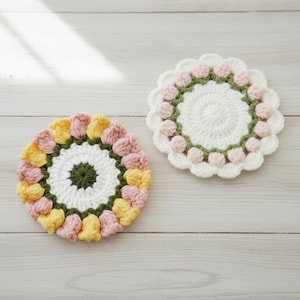 May include: Two handmade crochet coasters. One coaster features a white centre with a green border and pink flower-like accents. The other coaster has a white centre, a green border, and is surrounded by pink and yellow flower-like accents.