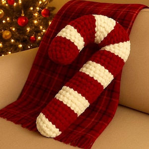 May include: A large, red and white striped candy cane-shaped cushion. The cushion is made of a chunky knit material and rests on a red plaid blanket. A festive holiday decoration.