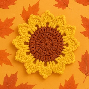 Puede incluir: Un posavasos de girasol tejido a ganchillo, con un centro marrón y pétalos amarillos. Se encuentra sobre un fondo naranja con hojas de otoño naranjas dispersas, creando un ambiente cálido y otoñal.