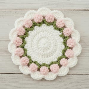May include: A handmade, white crochet coaster with a scalloped edge. The coaster features a ring of pink pom-poms and a green border, creating a floral design. The center is a solid white crochet circle.