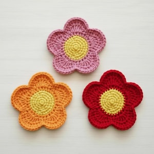 May include: Three handmade crochet flower coasters in shades of pink, orange, and red. Each coaster features a yellow center and petal design. The coasters are arranged on a white surface, showcasing their vibrant colors and textured details.