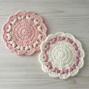 May include: Two handmade crochet coasters. One is light pink with white accents, and the other is white with pink accents. Both have a scalloped edge and a floral design. The coasters are circular and appear to be made of soft yarn.
