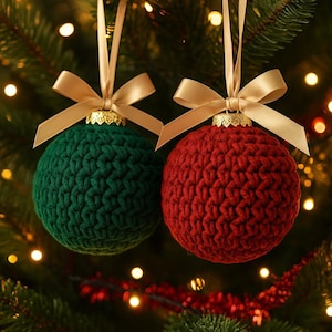 May include: Two crocheted Christmas ornaments. One is green, the other is red. Each sphere is topped with a gold-colored metal cap and a gold ribbon bow. The ornaments hang against a backdrop of a Christmas tree with twinkling lights.
