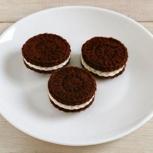 May include: Three crocheted cookies with a dark brown exterior and a white cream filling are arranged on a white plate. The cookies are round and have a textured appearance, resembling a popular sandwich cookie.