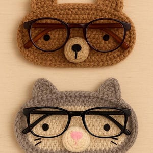 May include: Two handmade crochet eyeglass holders. One is brown and shaped like a bear, the other is gray and shaped like a cat. Each holder has a pair of eyeglasses resting on it. The bear holder has brown glasses, and the cat holder has black glasses.