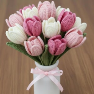 May include: A bouquet of handmade crochet tulips in shades of pink and white, with green leaves, arranged in a white vase tied with a pink ribbon. The flowers are crafted with detailed stitching, creating a textured appearance.
