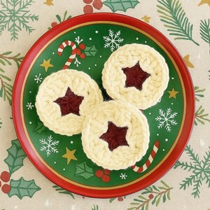 Könnte beinhalten: Ein roter und grüner Weihnachtsteller mit drei cremefarbenen, gehäkelten Keksen. Jeder Keks hat eine sternförmige, dunkelrote Mitte. Der Teller ist mit Zuckerstangen, Schneeflocken und Sternen verziert. Der Hintergrund ist ein festliches Stoffmuster.