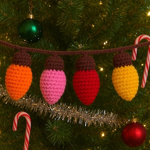 May include: A string of four crocheted Christmas light ornaments in orange, pink, red, and yellow with brown tops. The lights are strung on a brown crocheted cord and are hanging on a Christmas tree with candy canes and ornaments.