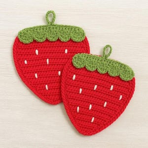 Może przedstawiać: Dwa ręcznie robione, czerwone, szydełkowe podkładki w kształcie truskawek z zielonymi wierzchami i białymi detalami nasion. Każda podkładka ma pętlę do zawieszenia. Podkładki mają chronić powierzchnie przed gorącem.